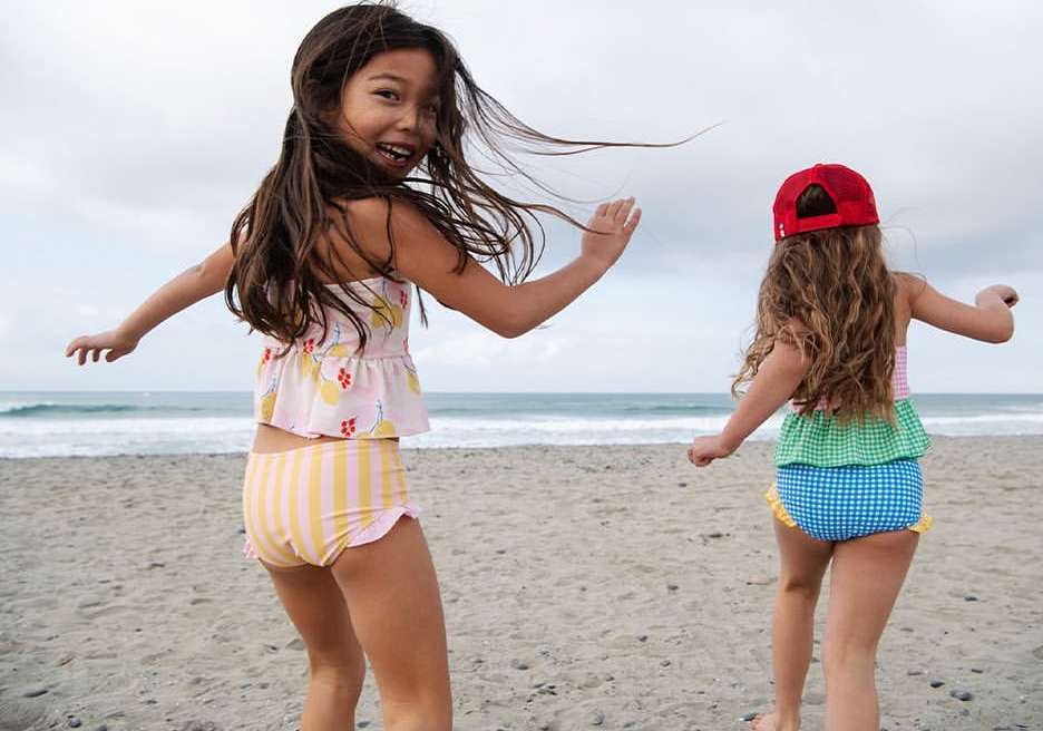 Two young girls in swimsuits play on a sandy beach near the ocean with a cloudy sky. One girl has long brown hair and is smiling.