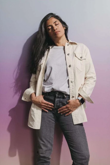 Adult female stands against a gradient background wearing casual clothing, jeans, and a buttoned jacket with her hands in her pockets.