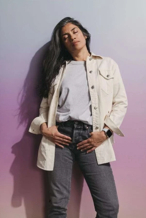 Adult female stands against a gradient background wearing casual clothing, jeans, and a buttoned jacket with her hands in her pockets.