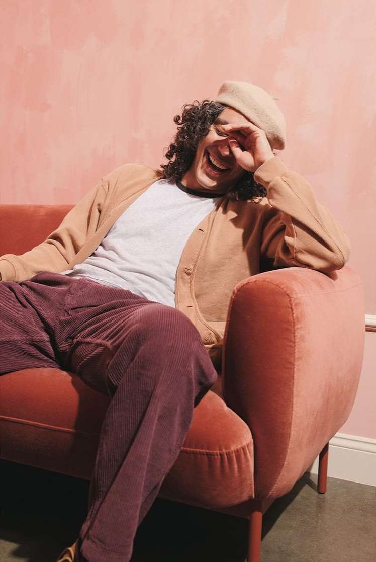 Man with curly hair laughs while lounging on a pink couch wearing a cardigan, t-shirt, beret, and corduroy pants in a casual indoor setting.