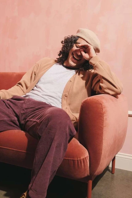 Man with curly hair laughs while lounging on a pink couch wearing a cardigan, t-shirt, beret, and corduroy pants in a casual indoor setting.
