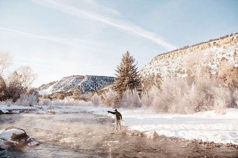 An adult male wearing a jacket and pants stands in a river in a snowy landscape, fly fishing with a fishing rod, mountain backdrop.