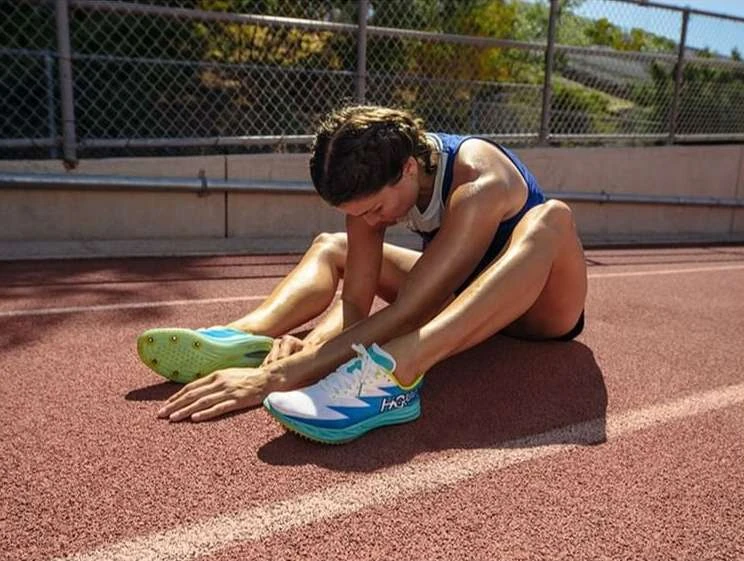 Female athlete in blue tank top sits on a red running track, stretching legs and wearing sports shoes, with trees in the background.