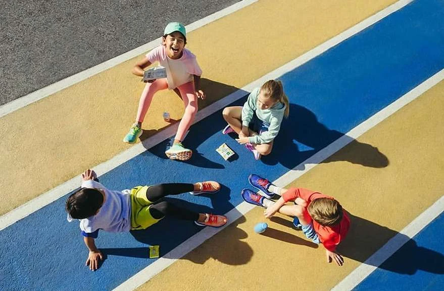 High-angle view of four children sitting on a multi-colored surface, snacking and interacting in bright sunlight with visible shadows.