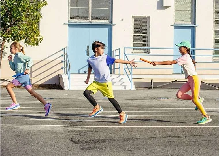 Three children are participating in a relay race outdoors on a running track near a building, passing a baton under bright daylight.