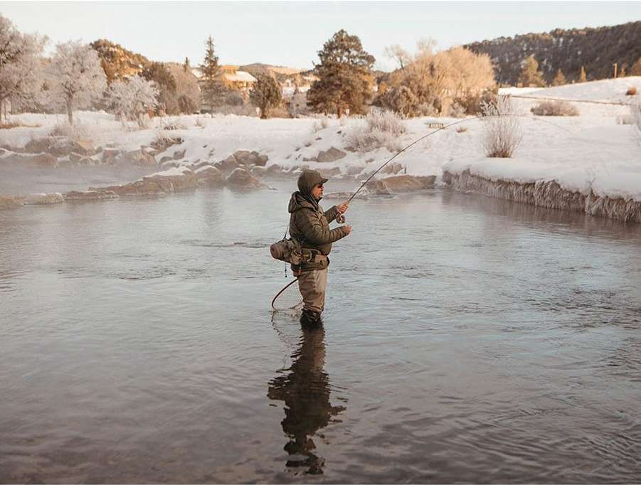 A man fly fishing in a cold river in winter, wearing waders and a jacket with snowy trees and a mountain in the background.