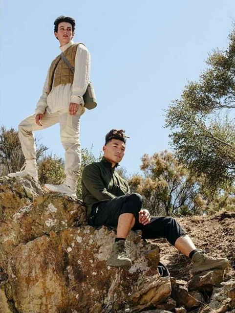 Two men pose on rocks in outdoor setting, wearing outdoor clothing and boots, against a backdrop of clear blue sky and trees.