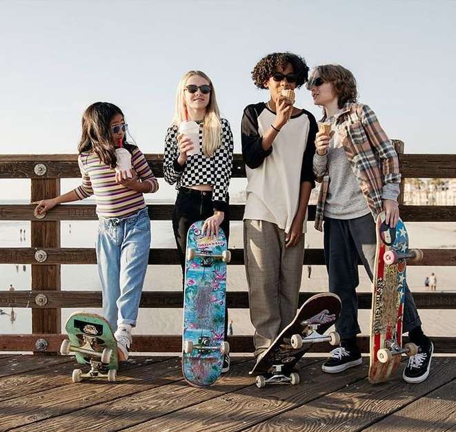 Four young people standing on a wooden pier holding skateboards and ice cream or smoothies with the ocean visible in the background.