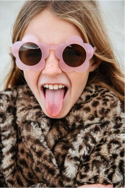 Young girl with flower sunglasses sticks out tongue, wearing animal print coat; playful expression, light background, portrait orientation.