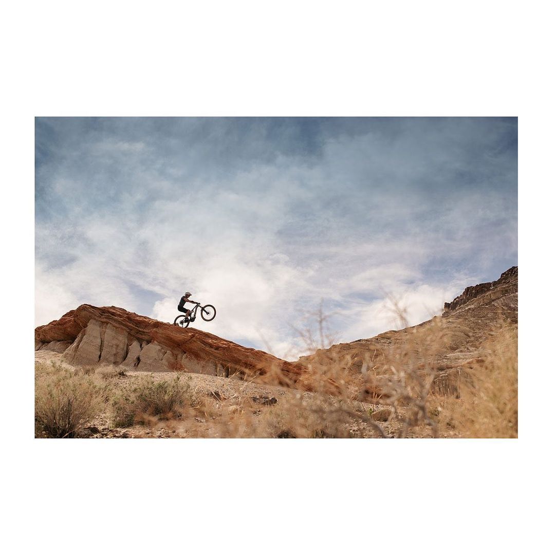 A person rides a mountain bike downhill on a red dirt track. The bike and rider are silhouetted against a cloudy sky and rugged terrain.