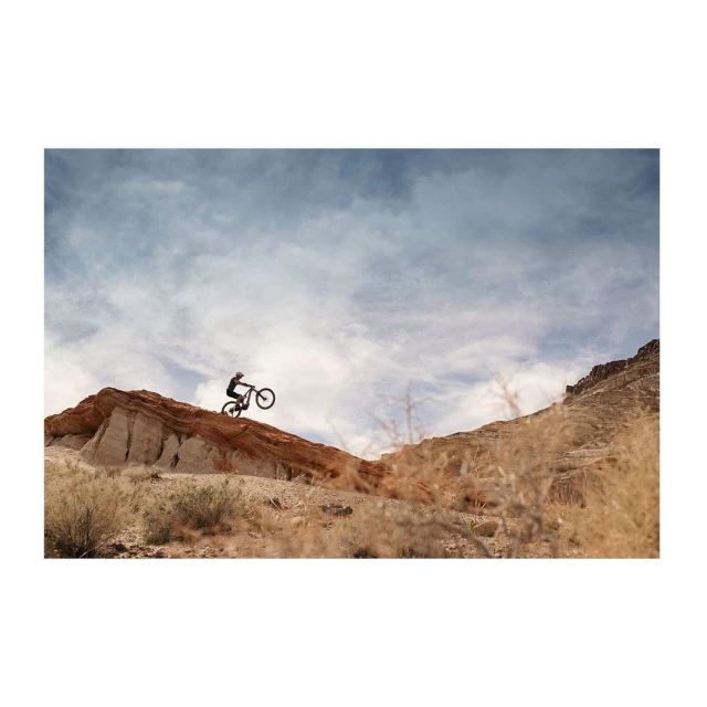 A person rides a mountain bike downhill on a red dirt track. The bike and rider are silhouetted against a cloudy sky and rugged terrain.