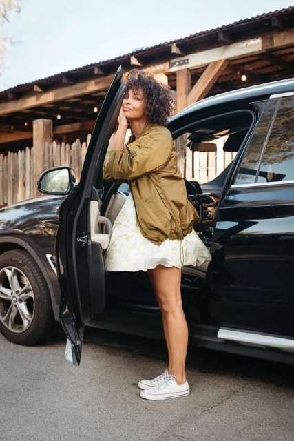 A woman exits a black SUV on a sunny day, wearing a white patterned dress and an olive-green jacket, standing on an asphalt surface.