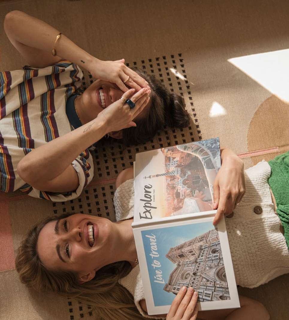 Two smiling women lying on a patterned rug with travel books featuring European landmarks, bathed in soft, diffused light indoors.