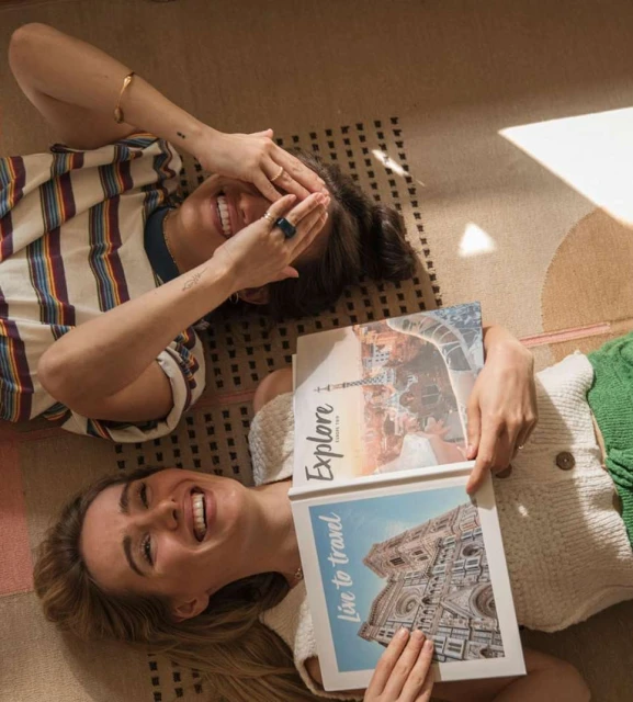 Two smiling women lying on a patterned rug with travel books featuring European landmarks, bathed in soft, diffused light indoors.