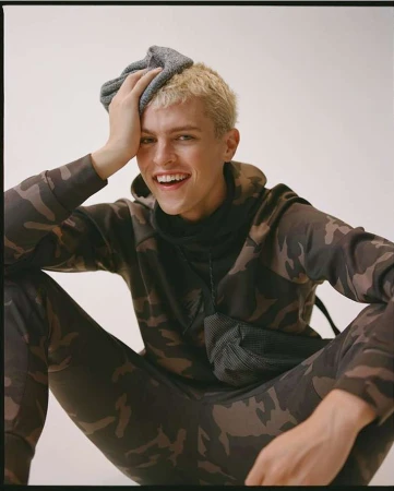 Smiling blonde man in a camouflage hoodie holding a grey beanie on his head against a white background.
