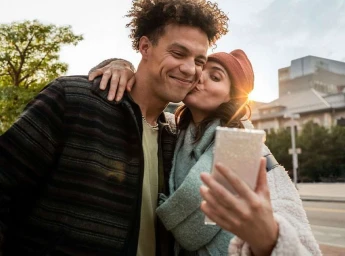 A young woman kissing a man on the cheek while holding up a smartphone for a selfie outdoors.