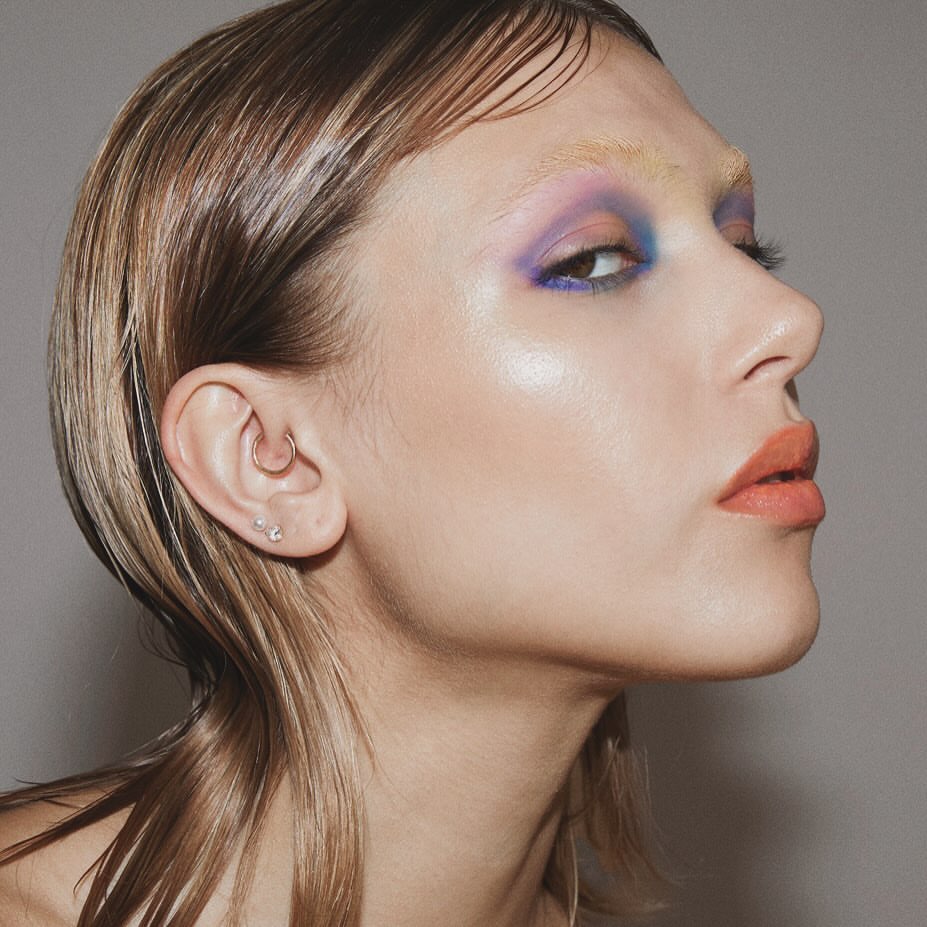 Close-up profile shot of a young adult female with blonde wet-look hair, colorful eye makeup, and coral lipstick against a grey background.