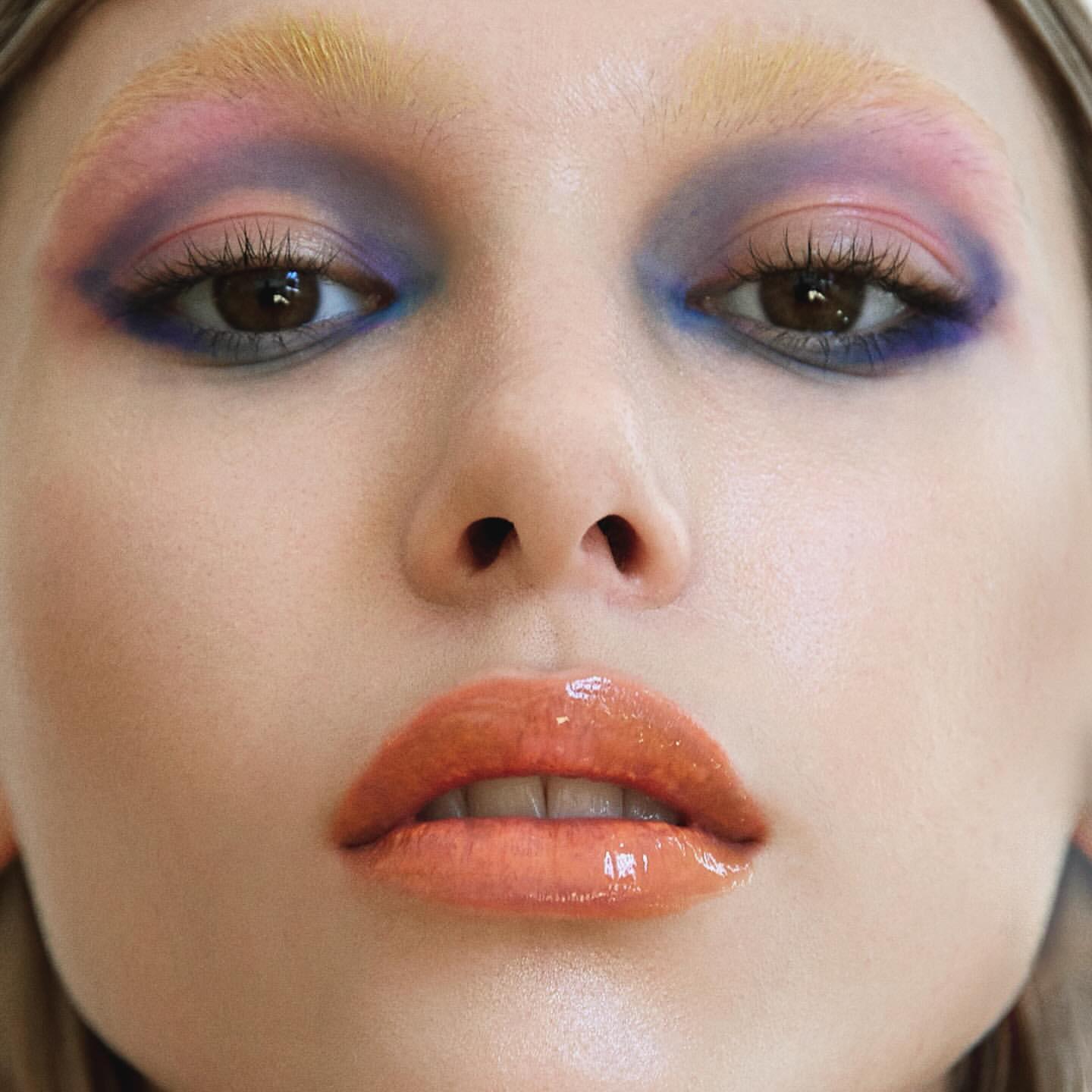 Close-up portrait of a woman with artistic makeup, featuring colorful eyeshadow, highlighted eyebrows, and glossy lips in a beauty shot.