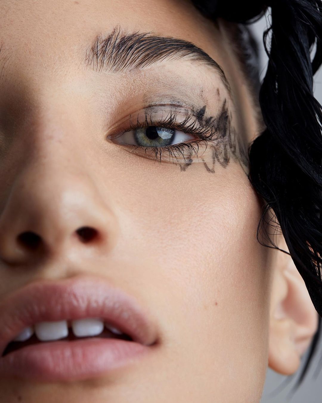 Close-up portrait of a woman with artistic black eye makeup and braided dark hair against a neutral background in a beauty shot.