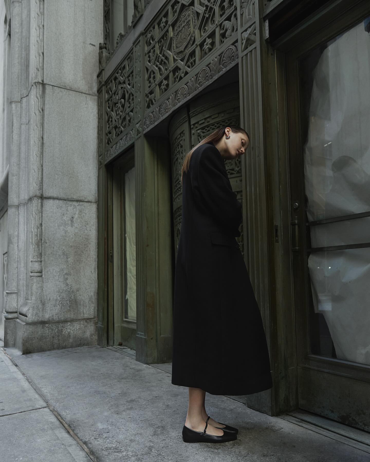 Woman in a black coat stands in front of an ornate building with classic architectural details on a cloudy day in an urban setting.