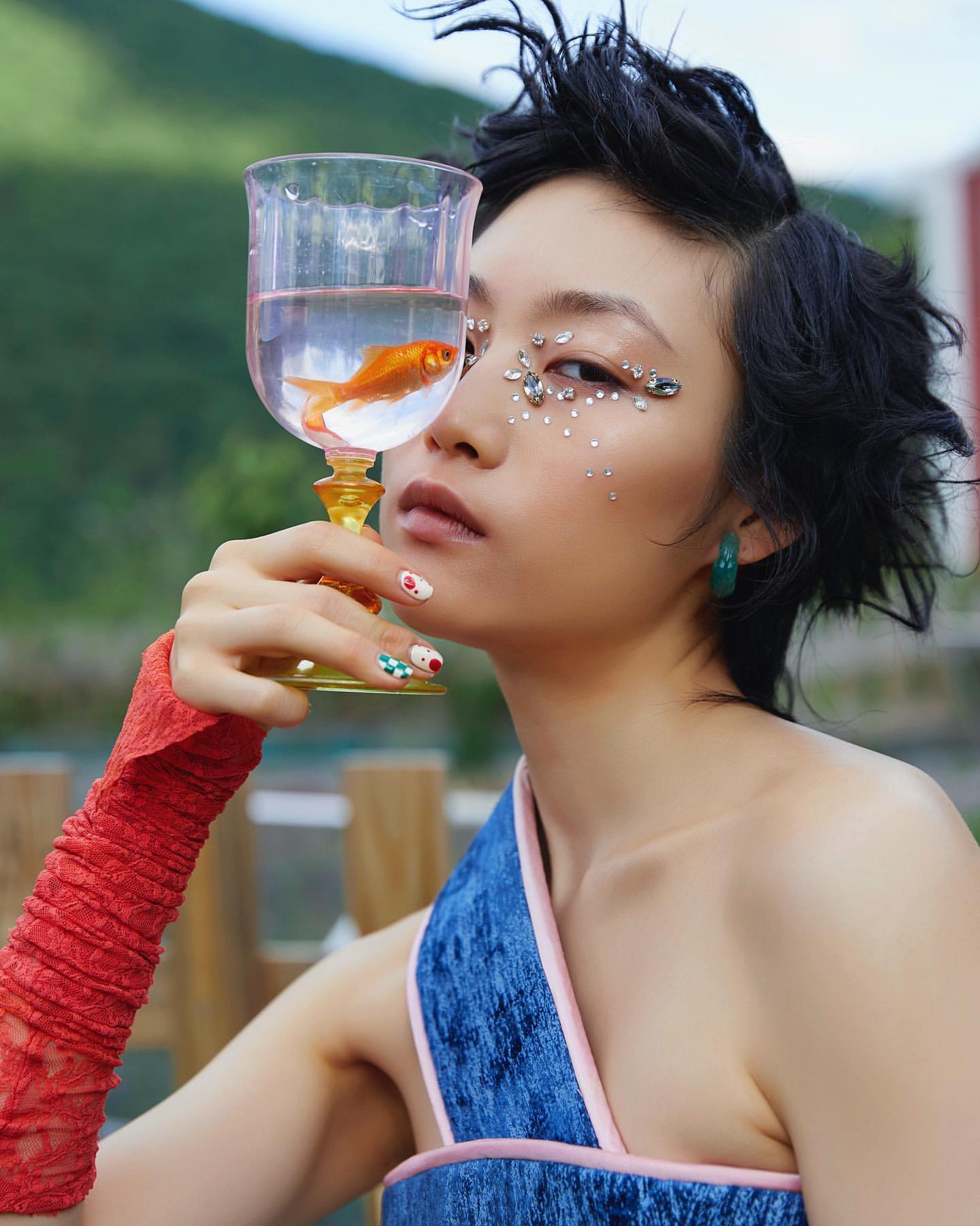 Woman poses with a glass holding goldfish in outdoor setting. She wears a blue dress, red glove, and face gems, creating a surreal look.