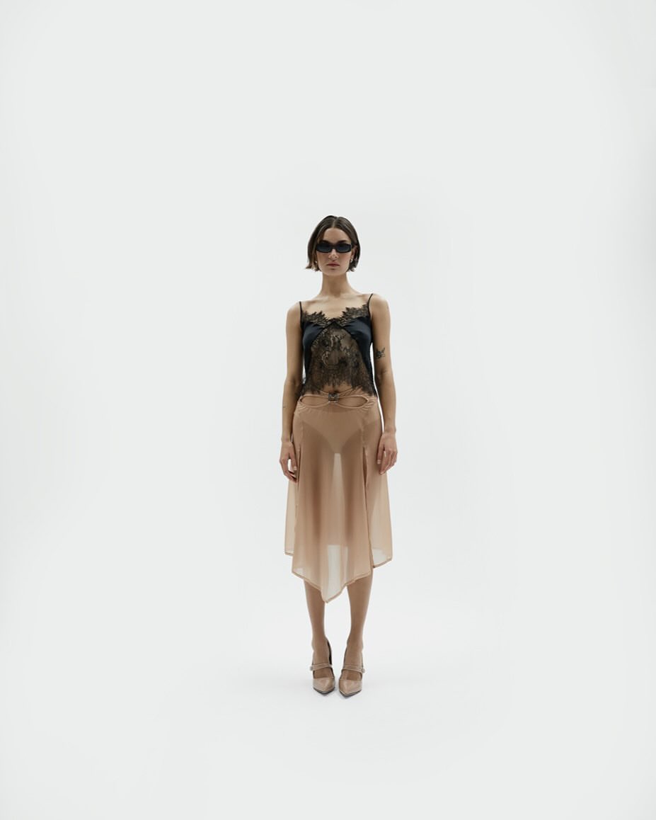 Full shot of a young woman with sunglasses posing in a black lace top and tan asymmetrical skirt in a bright white photo studio.