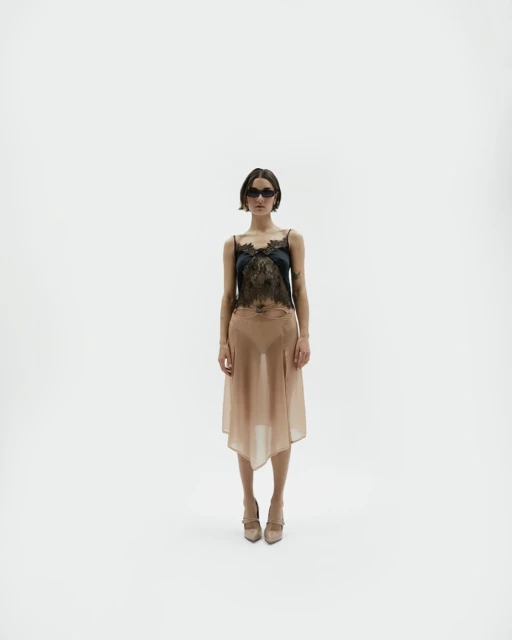 Full shot of a young woman with sunglasses posing in a black lace top and tan asymmetrical skirt in a bright white photo studio.