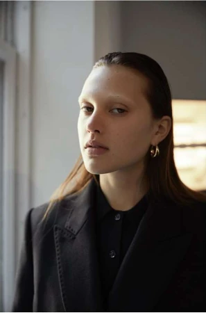 Portrait of a woman with slicked-back hair wearing a dark blazer and gold hoop earrings near a window