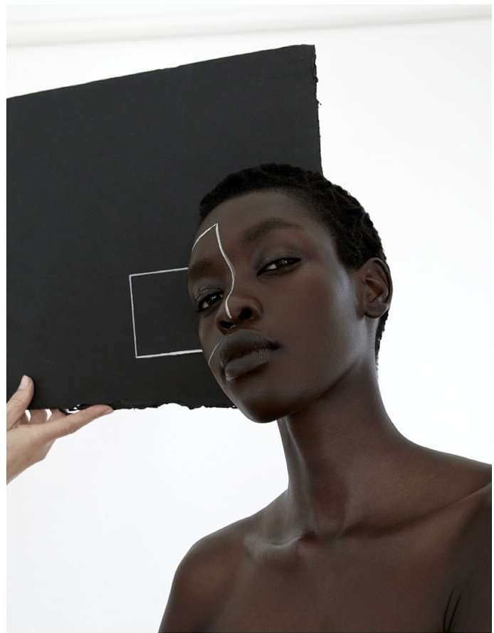 Headshot of a Black woman with white graphic face makeup, next to a held black square against a white backdrop in a photo studio.