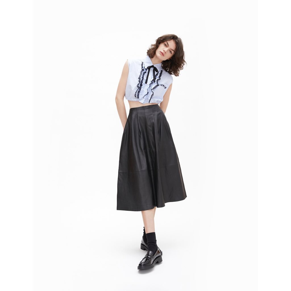 A slender woman in a light blue blouse with black ribbon detail and a black midi skirt poses in a full shot on a white background.