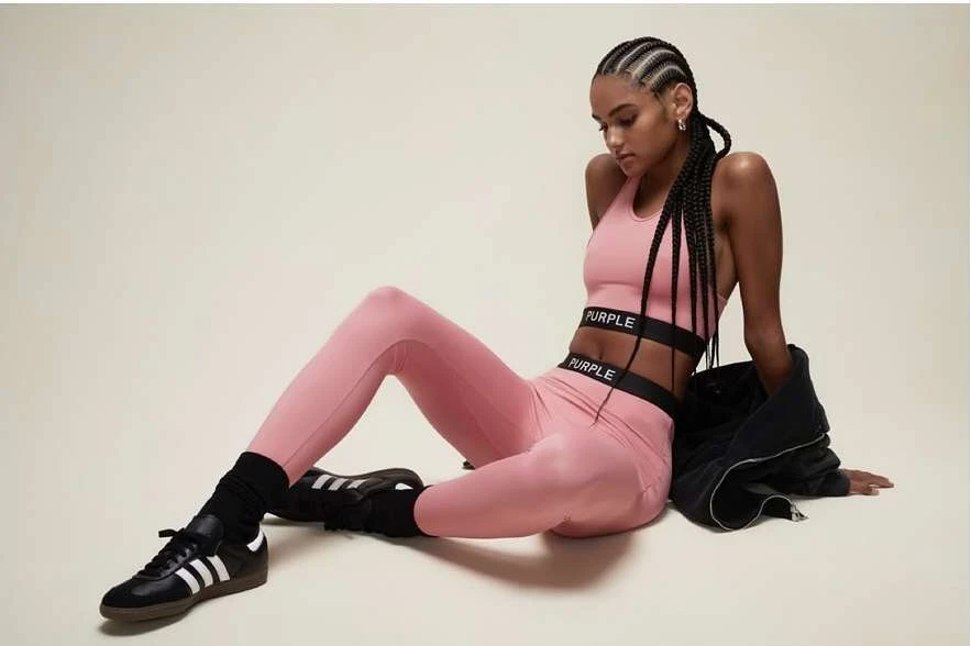 A young adult woman with braided hair poses in pink athletic apparel, wearing black and white Adidas sneakers against a neutral backdrop.