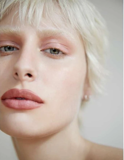 Close-up portrait of a young adult woman with short blonde hair, wearing pink eyeshadow and lipstick against a neutral background.