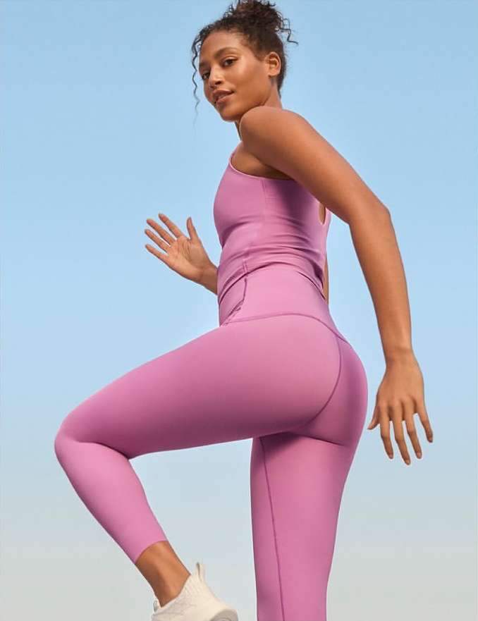 A woman in purple athletic wear confidently strides against a pale blue sky, showcasing fitness and sportswear fashion in a dynamic pose.