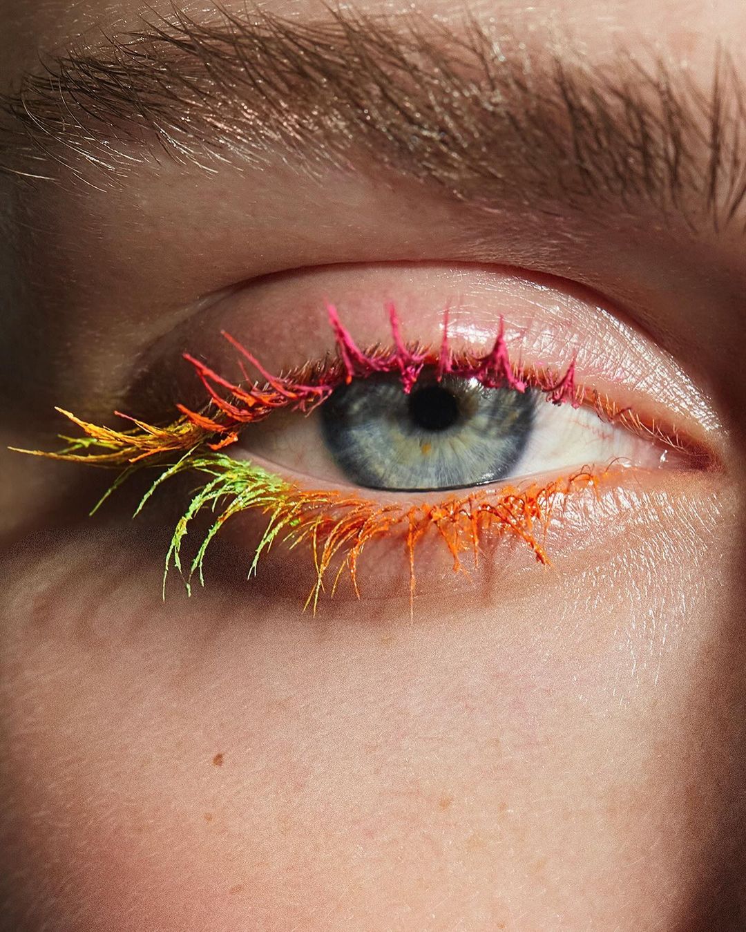 Close-up of a blue eye with vibrant neon pink, orange, and yellow mascara on the eyelashes for a striking beauty statement.