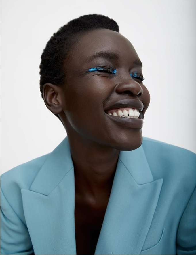 Close-up portrait of a smiling African-American woman in a light blue suit with teal eyeshadow against a plain white backdrop.