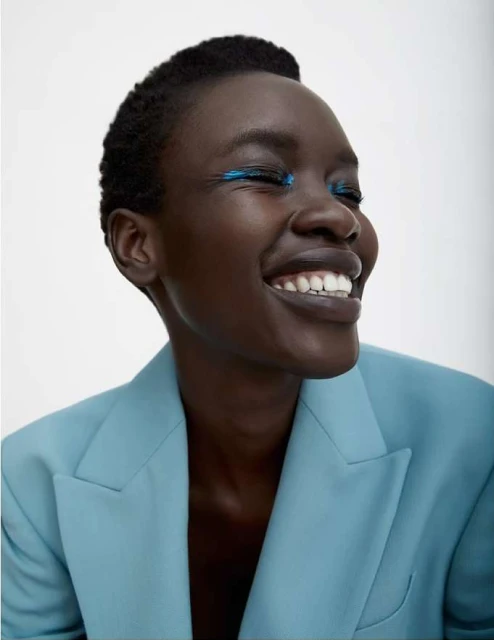 Close-up portrait of a smiling African-American woman in a light blue suit with teal eyeshadow against a plain white backdrop.