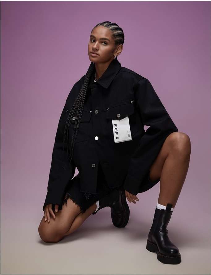 Young woman in black denim jacket and shorts poses kneeling in studio with purple background; fashion-forward style and confident expression.