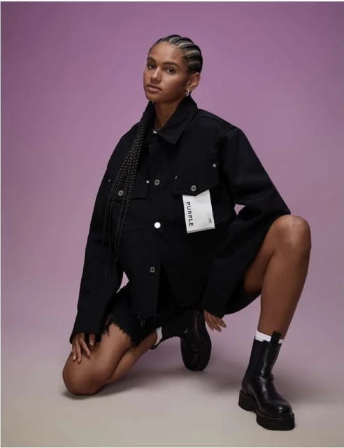 Young woman in black denim jacket and shorts poses kneeling in studio with purple background; fashion-forward style and confident expression.