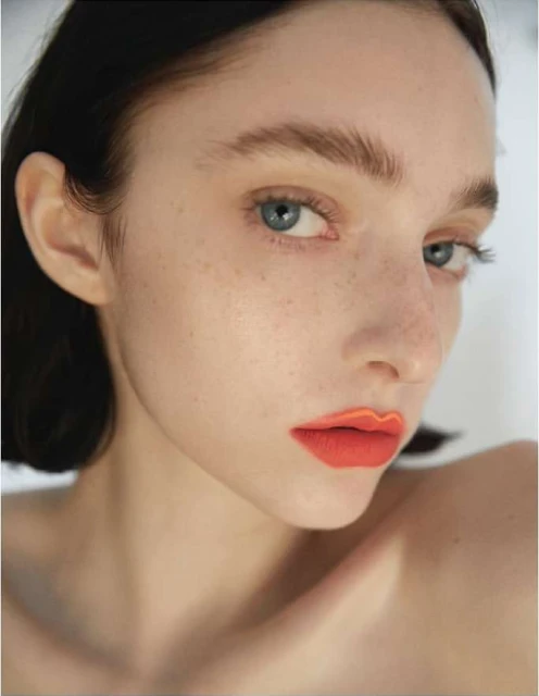 Close-up of an adult female model with fair skin and freckles, wearing orange lip color, posing against a white backdrop in a studio setting.