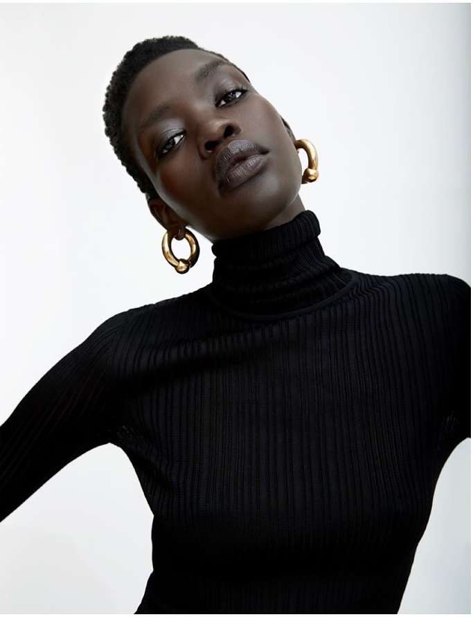 Headshot of an African-descent woman in a black turtleneck sweater and gold hoop earrings against a neutral background in soft light.