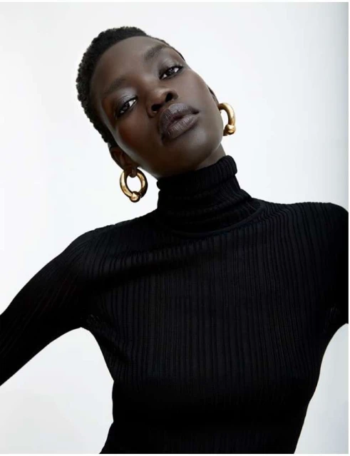 Headshot of an African-descent woman in a black turtleneck sweater and gold hoop earrings against a neutral background in soft light.