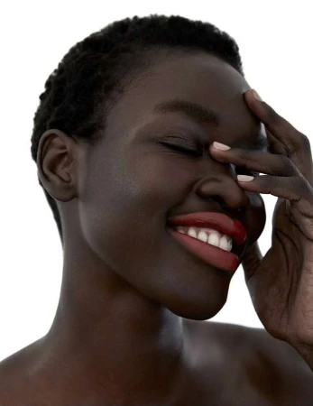 Close-up portrait of a smiling Black woman with short dark hair wearing deep red lipstick, touching her forehead against a white background.