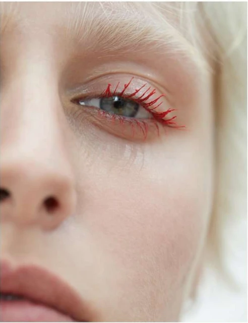 Extreme close-up of a fair-skinned person's face, featuring red mascara on the upper lashes and light-colored hair against a bright backdrop.