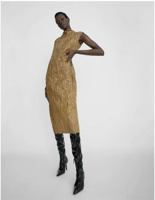 Fashion model poses in textured gold dress and high-heeled black patent leather boots against a white backdrop in a fashion shot.