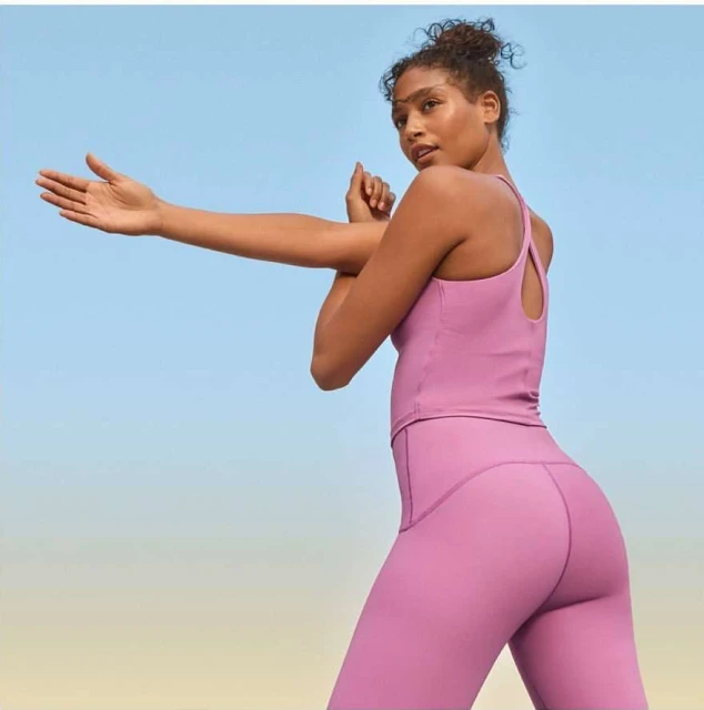 Athletic woman in pink workout clothes stretches her arms in front of a blue sky, demonstrating active lifestyle and fitness routine.