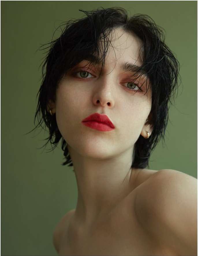 Close-up portrait of a woman with short dark wet hair, red lipstick, and an olive green background.