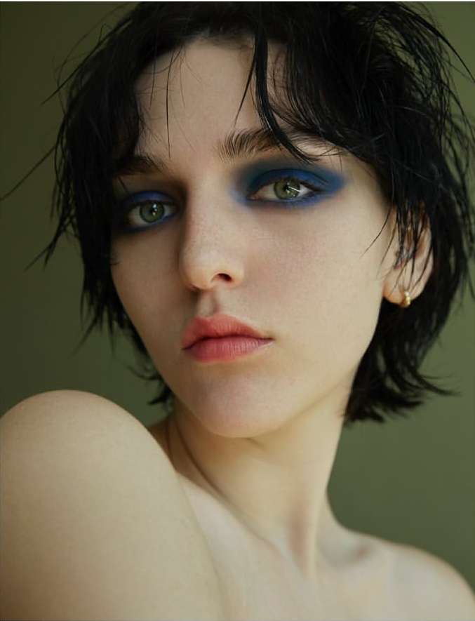 Close-up portrait of a young adult woman with dramatic blue eyeshadow and short, dark hair against a solid green background.