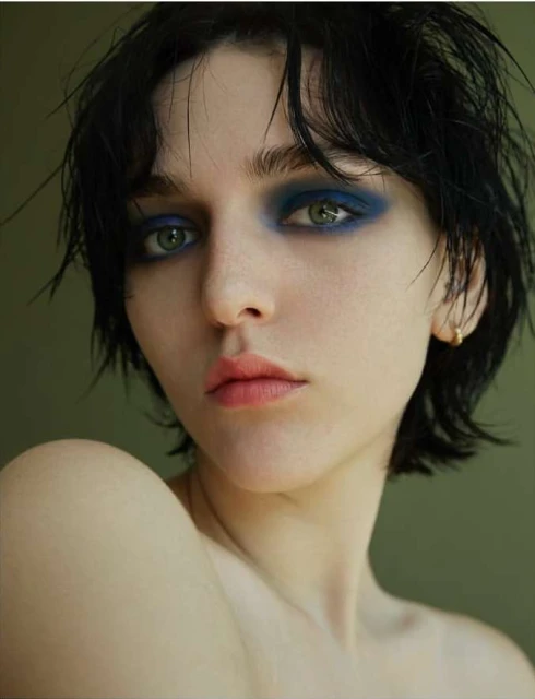Close-up portrait of a young adult woman with dramatic blue eyeshadow and short, dark hair against a solid green background.