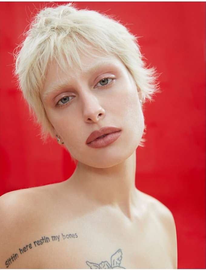 Close-up portrait of a woman with short blonde hair and visible tattoos against a bright red background.