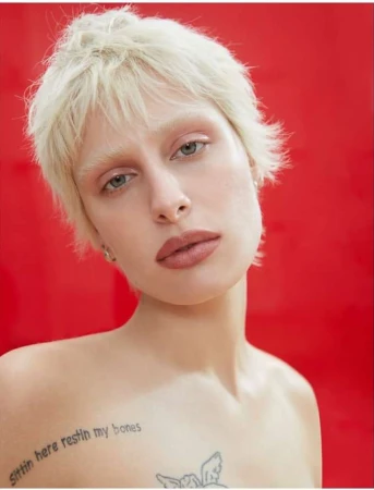 Close-up portrait of a woman with short blonde hair and visible tattoos against a bright red background.